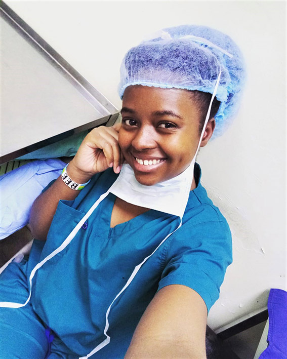 На фото — медсестра из Руанды Бахати в синей одежде медсестры с сеткой для волос, с маской для защиты лица, спущенной на шею, во время рабочего перерыва. Она улыбается для селфи.