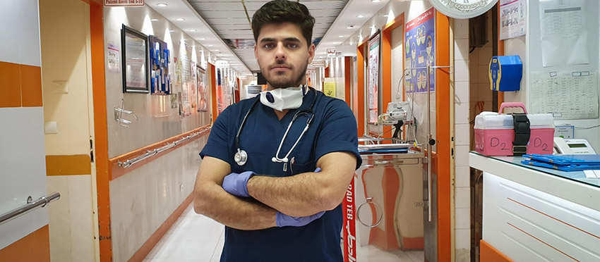 Young refugee Moheyman Alkhatavi can be seen in the hallway of a hospital where he works as a nurse treating COVID-19 patients. He wears the official workwear of a nurse, and has a face mask and stethoscope hanging around his neck.