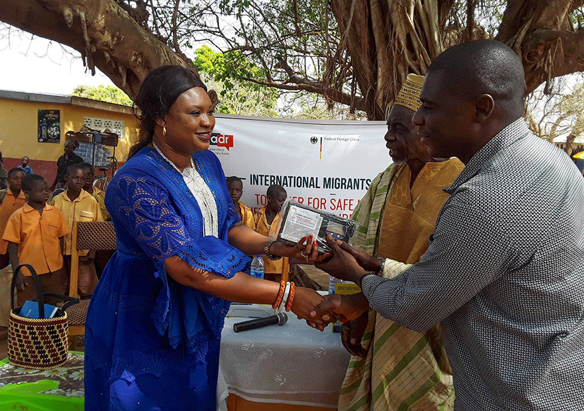 WADR station manager Agnes Thomasi is standing on the left, greeting two community members in Ghana at a ceremony on International Migrants Day. As a gift, Agnes Thomasi hands over radio equipment.