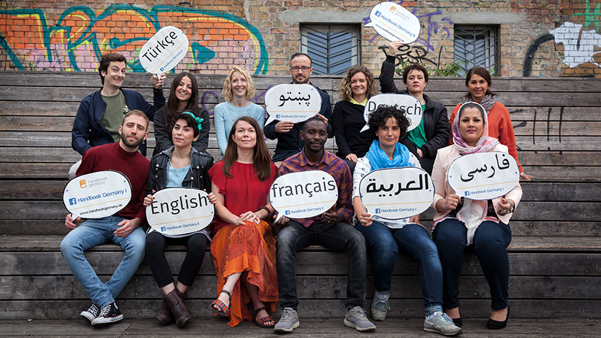 13 members of the multilingual Handbookgermany.de editorial team sitting on a public bench holding up speech bubble posters indicating the seven languages that can be found on their information platform. The languages are German, English, Arabic and Farsi, as well as French, Pashto and Turkish.