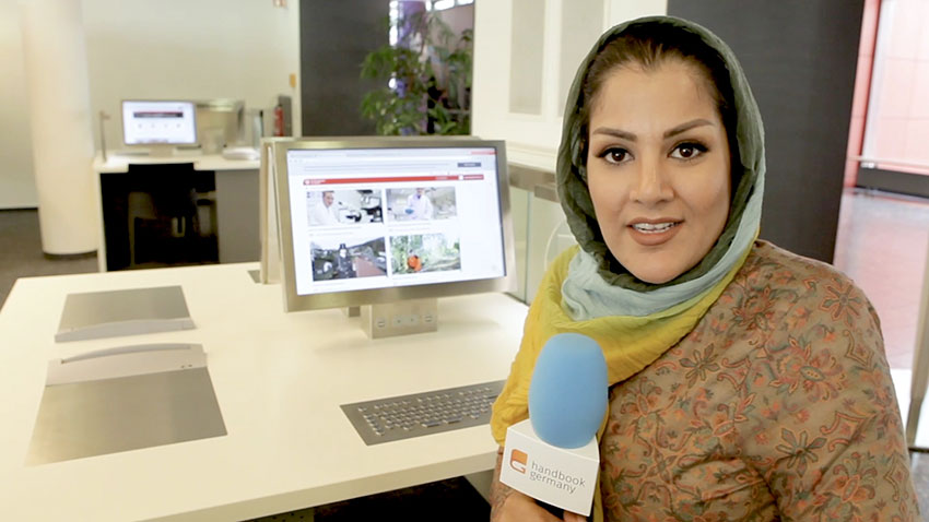 Reporter Sharmila Hashimi from Handbookgermany.de sitting in her office holding a microphone in front of her desktop computer in preparation for a show.