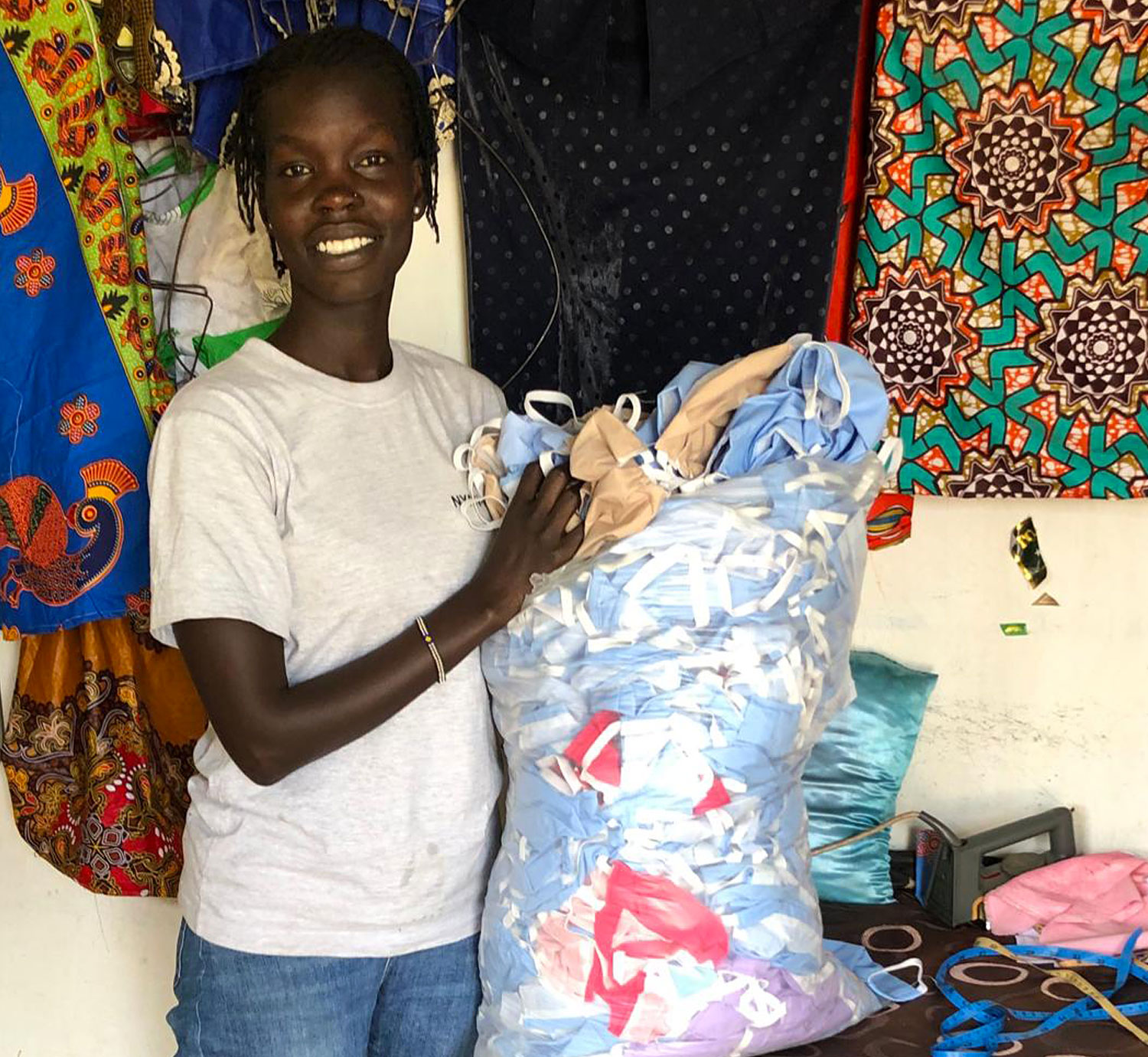 A young woman called Adhieu who lives in Kenya is seen kneeling on the ground and producing self-made soap by filling the mixed pre-products into forms that can be divided into individual pieces of soap. The young woman is also seen with a bag full of self-made face masks in various colours.