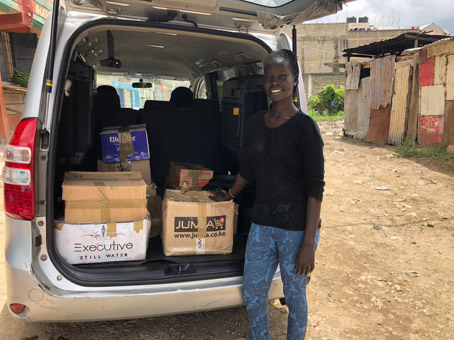A young woman named Adhieu stands by a minivan with the tail gate open. In the van are cardboard boxes filled with self-made face masks that will be distributed in a refugee camp.