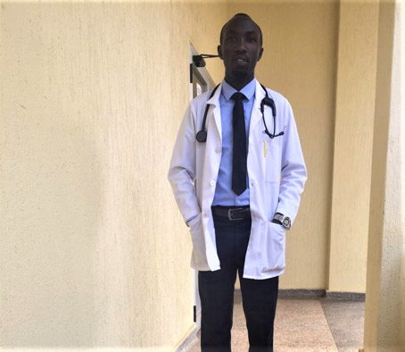 Sur la photo, on peut voir le « Dr Jonas », jeune médecin congolais, dans un couloir d’hôpital vide en tenue vestimentaire blanche de médecin, un stéthoscope au cou.